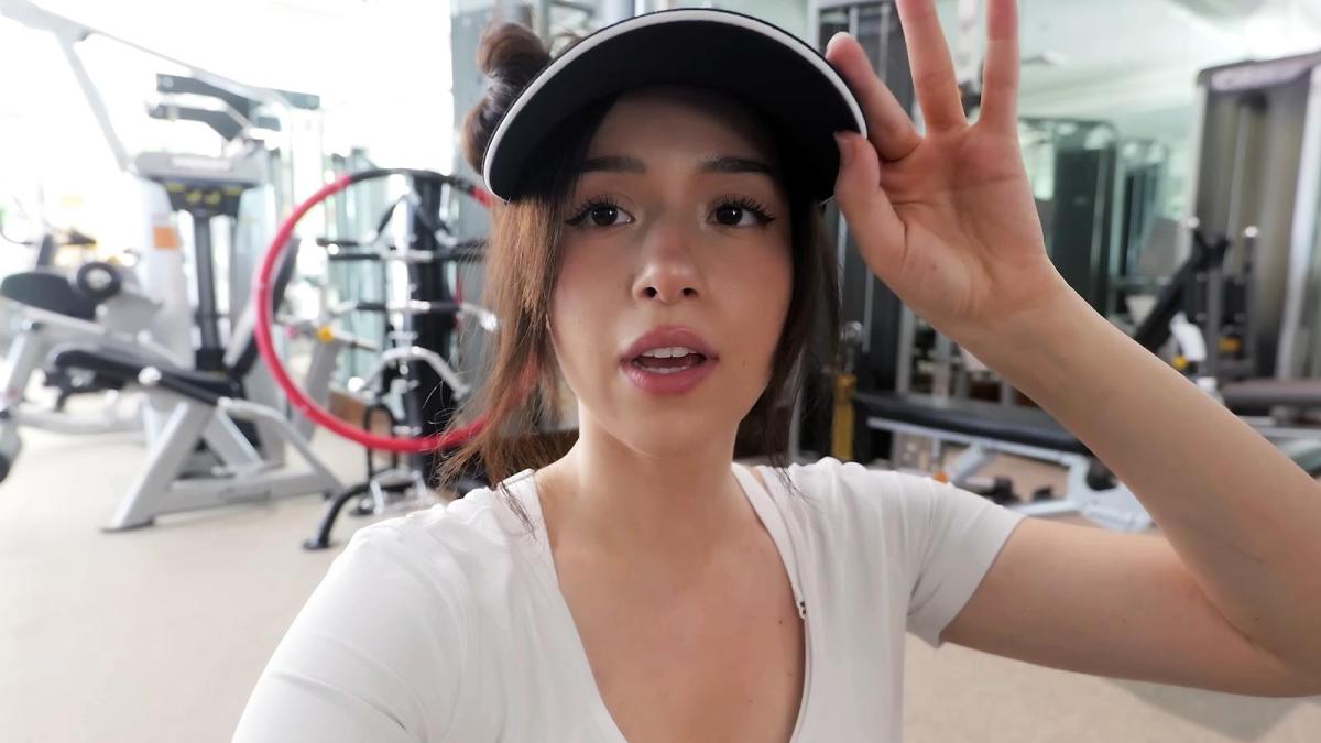 Pokimane holds her cap with her left hand while speaking to her streaming fans while working out at the gym.